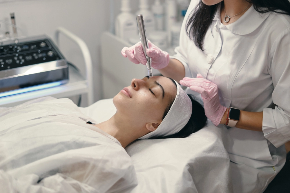 Skincare professional in gloves performs a facial treatment on a patient lying with eyes closed, using a handheld device.”