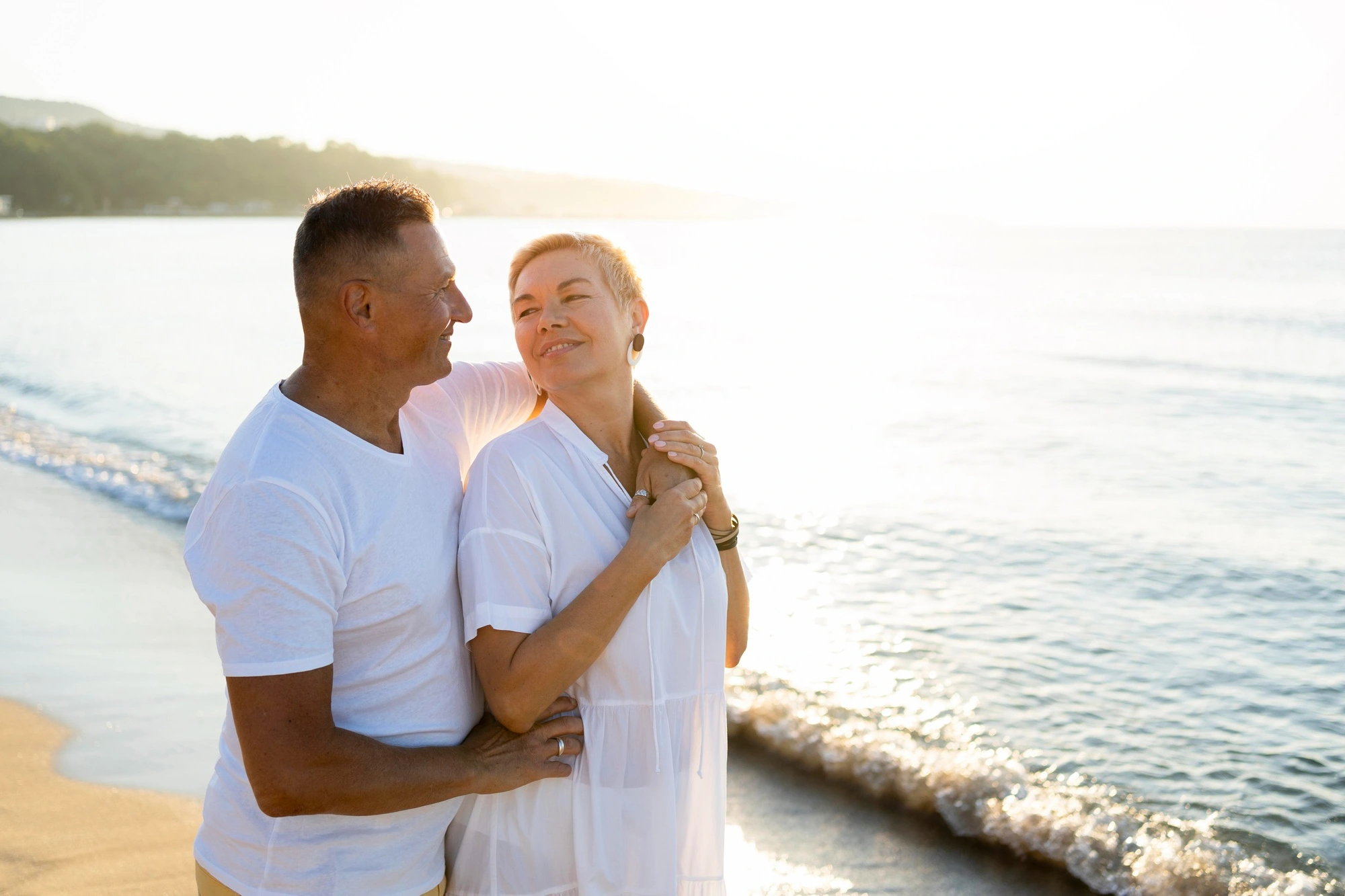 Couple embracing at the beach, smiling at each other with the ocean and sunlight behind them.