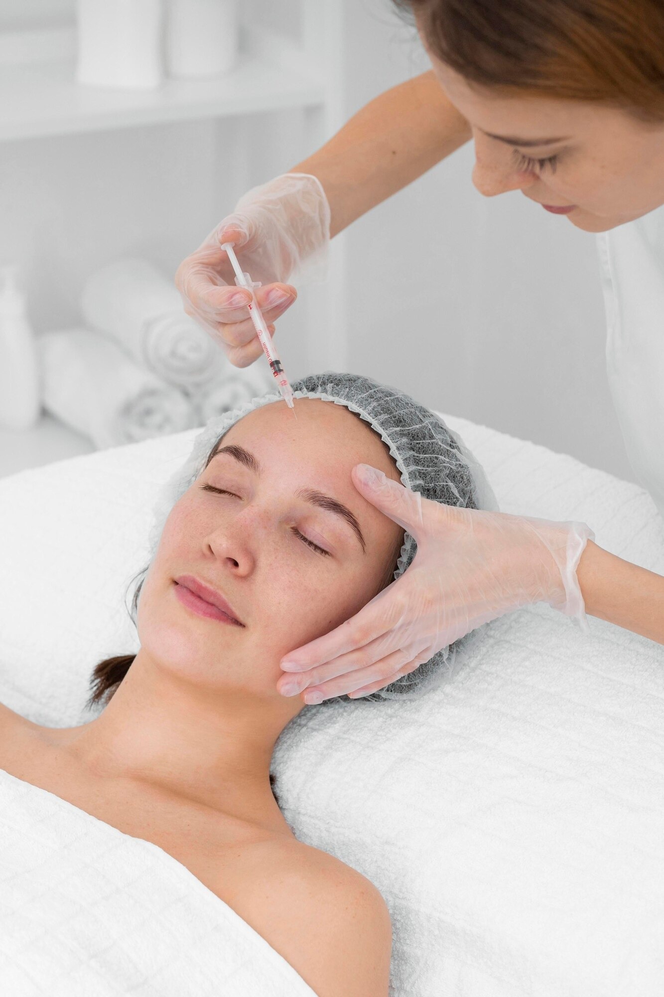 Medical aesthetic procedure: a nurse injects a patient’s forehead with a syringe while the patient lies relaxed on a treatment bed, wearing a disposable cap and gloves.