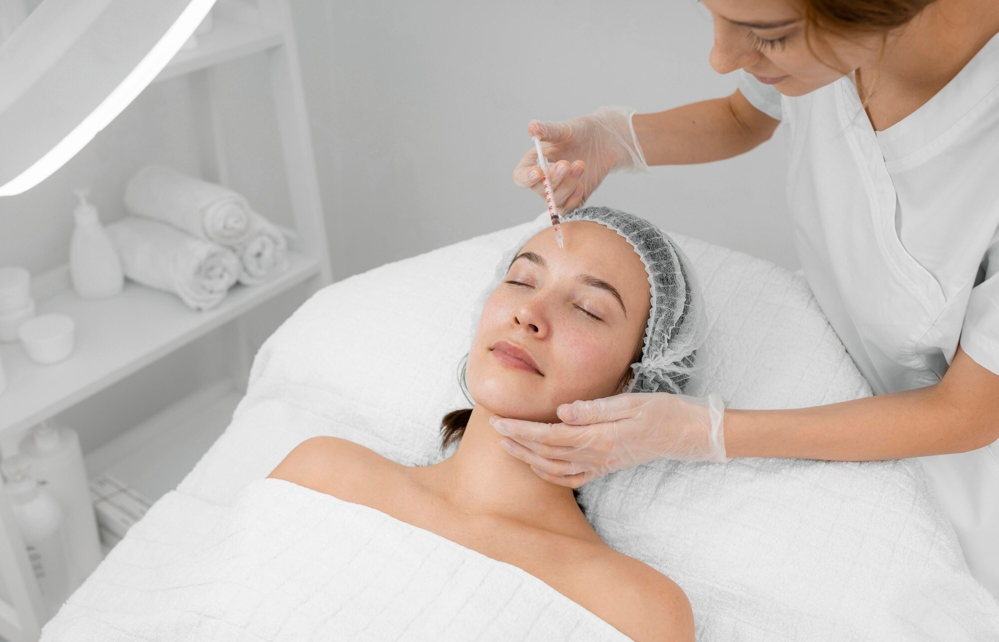 Esthetician administers a cosmetic forehead injection to a client who is lying with eyes closed in a spa setting.