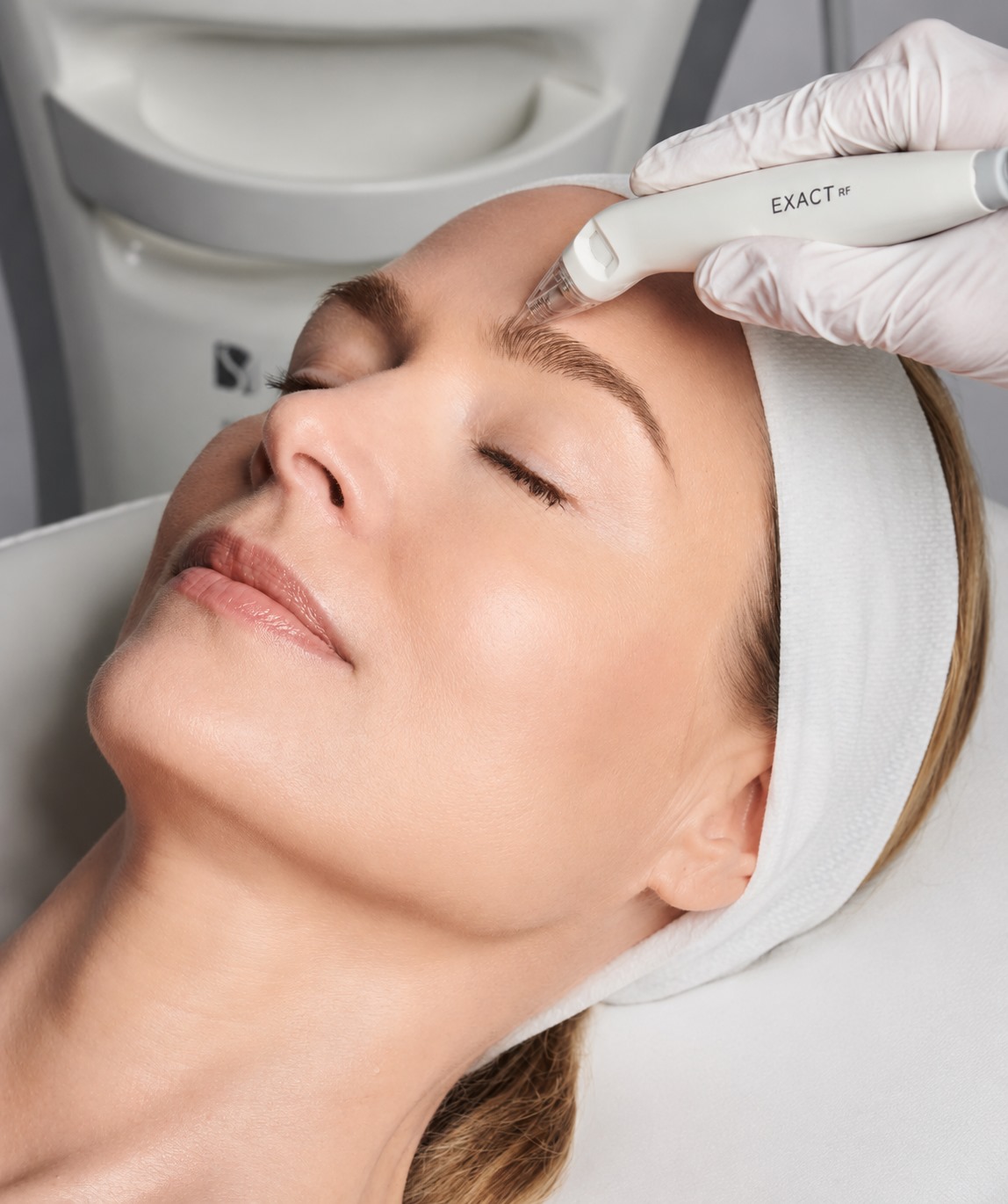 Woman receiving RF skin-tightening treatment near her eyebrow with a clinician wearing gloves and a white headband.