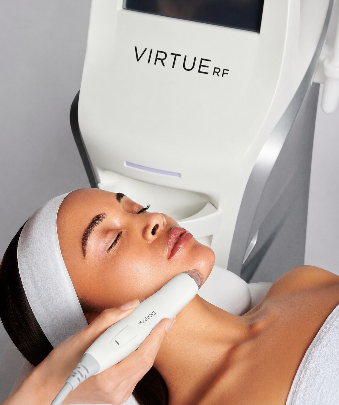 Woman lying down with eyes closed as a clinician uses a handheld Virtue RF device on her face/neck area during a skincare treatment.