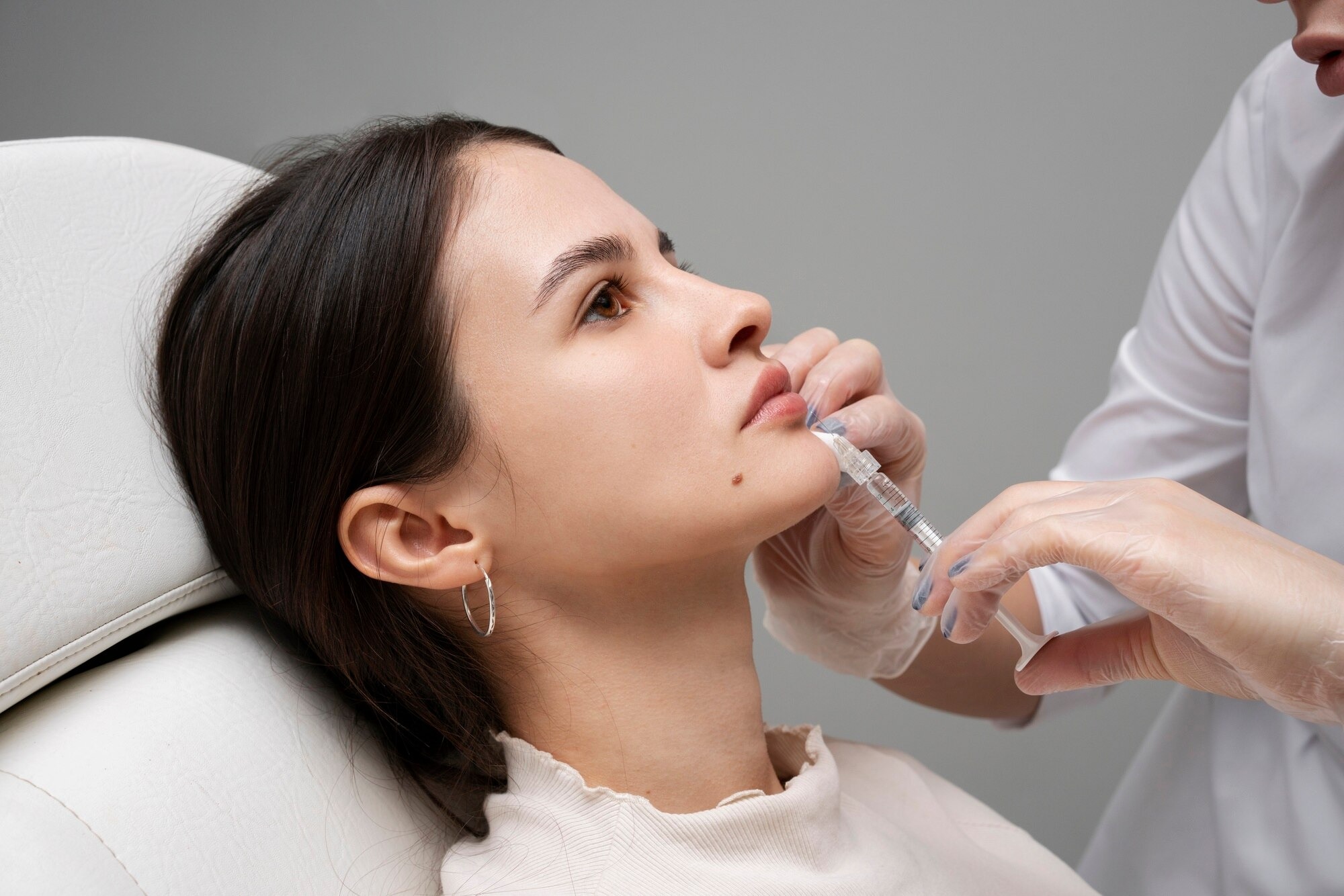 Woman lying in a chair while a clinician administers an injection to her lip area with a syringe and gloved hands.