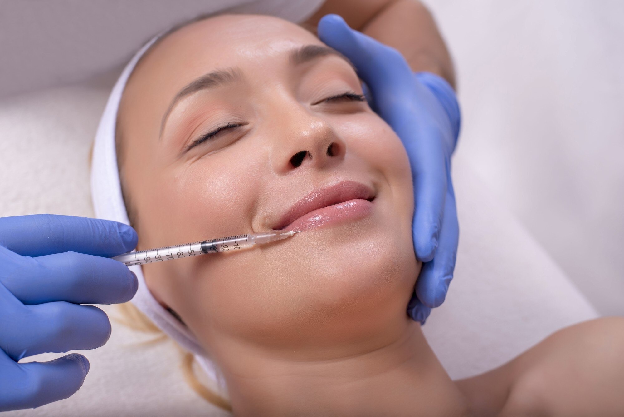 Clinician with blue gloves injects a patient’s lips for cosmetic filler, patient eyes closed and relaxed.