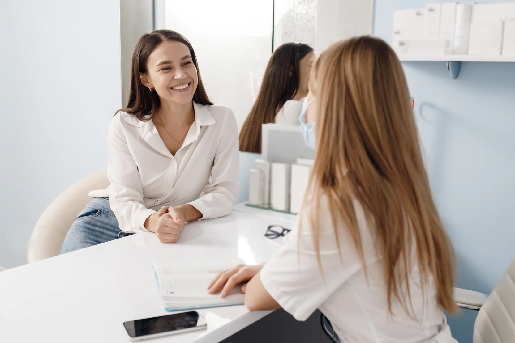 Smiling woman in a white blouse sits at a white desk across from a staff member in a clinic setting.