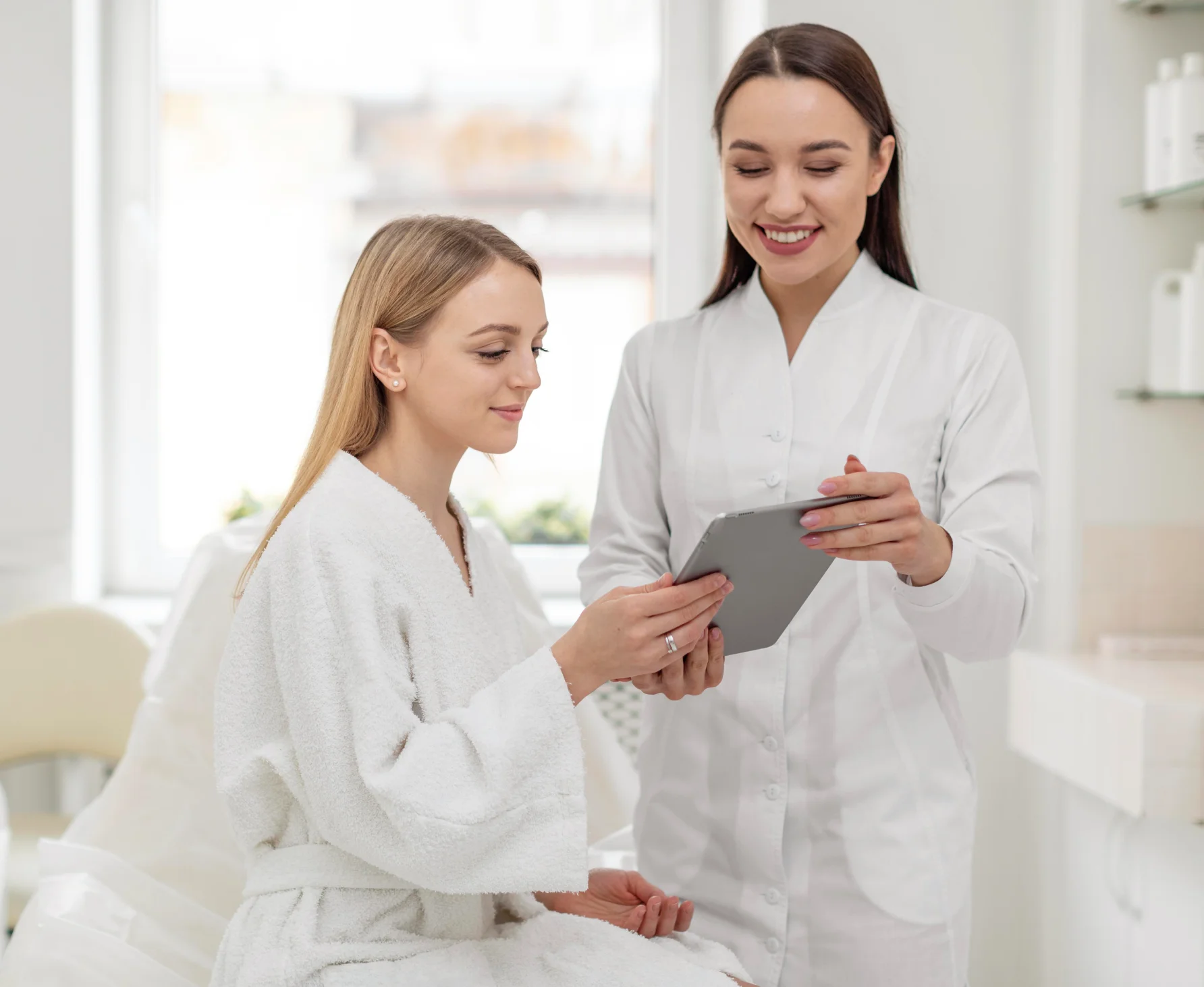 Clinician in a white uniform shows a tablet to a smiling patient in a white robe inside a bright clinic.