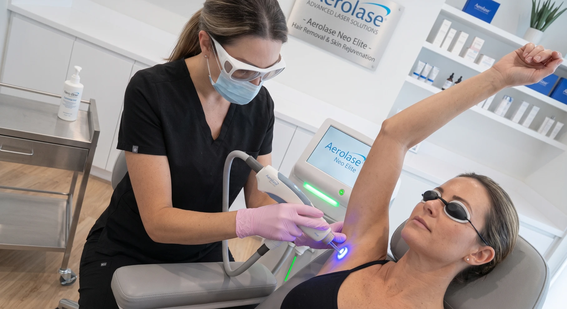 Clinician in PPE using a laser device on a patient’s raised armpit for hair removal (Aerolase Neo Elite) in a bright clinic.