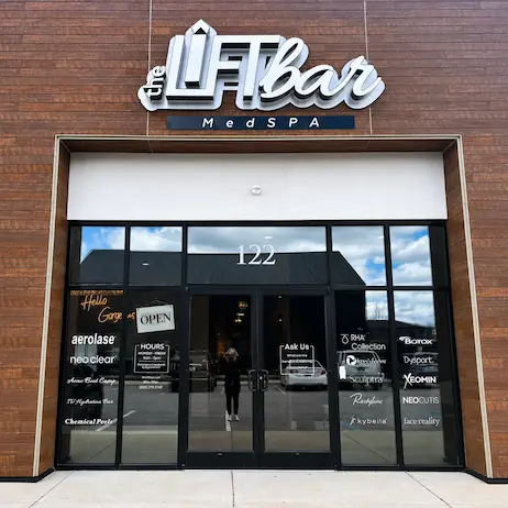 Storefront of a MedSpa with glass doors and dark metal frame, brick exterior, sign reading 'the LFT Bar MedSpa' above the entrance, street number 122 on the door panes, reflections of the parking lot in the windows.