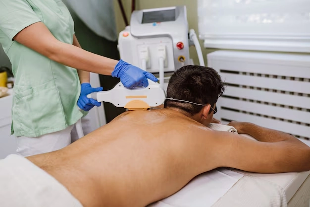 A clinician in green scrubs and blue gloves performing a cosmetic laser treatment, such as hair removal, on a man's upper back.